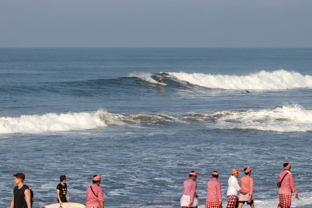 Surfing doing a cutback as locals hold a ceremony on the beach.